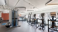 This fitness center is a comprehensively equipped, single-floor facility offering a generous workout space designed to accommodate a wide range of exercise disciplines. The layout is organized into distinct functional zones, maximizing the usability of the floor area.

The cardio section runs prominently along one side of the space, lined with a row of treadmills, elliptical trainers, and stair climbers. Large windows positioned along this wall flood the area with natural light and provide outdoor views, creating an open and energizing atmosphere during workouts. Rubber flooring throughout the facility provides durability and safety underfoot.

A dedicated strength and functional training zone occupies a separate area, featuring a Matrix cable machine, rowing machines, stationary and spin bikes, and adjustable benches paired with free weights. A large wall mirror in this section is framed by a terracotta accent wall, adding visual depth and aiding form during training.

The ceiling features exposed conduit, ductwork, and industrial-style fluorescent lighting fixtures, giving the space a utilitarian yet clean aesthetic. Structural columns divide portions of the floor plan naturally, while a wall-mounted flat-screen TV serves as a focal point. The overall ambiance is bright, functional, and purposefully arranged to support diverse fitness routines.