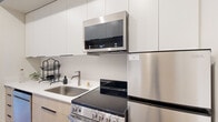 This efficiently designed one-bedroom, one-bathroom unit is laid out across a single floor, making smart use of every square foot. The layout flows from a central hallway — equipped with built-in open shelving and wall hooks for storage — into the main living areas. Light wood flooring runs throughout, creating a cohesive and warm aesthetic.

The kitchen is outfitted with a full suite of stainless steel appliances, including an electric range, over-the-range microwave, refrigerator, and dishwasher. White upper cabinetry contrasts with light wood lower cabinets, and clean white countertops provide a contemporary finish. The kitchen is compact yet fully functional.

The living room benefits from a large window and a glass-panel door that allows natural light to filter in and provides access to the outdoors. The neutral wall tones and light flooring contribute to an open, airy feel in the space.

The bedroom is a cozy, enclosed nook featuring a built-in wall shelf above the bed area, ideal for display or storage. The bathroom includes a white undermount sink set in a light wood vanity with a quartz-style countertop, a large mirror, and a clean, neutral color palette that keeps the space feeling fresh and orderly. Together, these rooms form a practical and modern living environment.