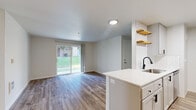 This well-appointed single-floor unit offers a clean, contemporary design throughout. The open-concept layout connects the kitchen and living area seamlessly, creating a spacious and airy feel. Large sliding glass doors in the living area flood the space with natural light and provide direct access to an outdoor green area, enhancing the sense of openness.

The galley-style kitchen is a standout feature, outfitted with white shaker cabinetry, matte black hardware, white quartz countertops, a white subway tile backsplash, and stainless steel appliances including a refrigerator, electric range, and over-range microwave. A deep single-basin sink with a matte black faucet and open wooden floating shelves add modern character to the space.

The living area extends comfortably from the kitchen, anchored by durable wood-look plank flooring that runs throughout the unit. Additional rooms offer bright, neutral walls, wood-look flooring, and ample natural light — one of which also features a sliding glass door with outdoor views. A wall-mounted heater unit provides climate control. The overall palette of soft whites and warm wood tones creates a cohesive, inviting ambiance suited to a variety of living arrangements.