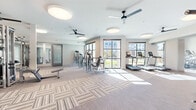 This well-appointed fitness center offers a generously sized, single-floor layout designed to accommodate a full spectrum of workout needs. The space is characterized by a clean, contemporary aesthetic, featuring neutral-toned patterned carpet tiles throughout that lend both visual interest and comfort underfoot.

Large floor-to-ceiling windows line one side of the facility, flooding the interior with abundant natural light and providing open views of the surrounding outdoor environment. The bright, airy atmosphere is further enhanced by high ceilings fitted with multiple modern circular flush-mount light fixtures and sleek black ceiling fans, ensuring excellent air circulation throughout.

The main floor is organized into distinct functional zones. A dedicated cardio area features a row of treadmills positioned directly in front of the windows, making for an engaging workout experience. The strength training section is equipped with a comprehensive selection of cable and resistance machines, free-weight benches, and adjustable equipment to support a wide range of exercises. A separate alcove area houses a TKO-branded functional training rig stocked with kettlebells, medicine balls, resistance bands, and a stability ball, complemented by a full-length mirror wall that enhances the sense of space. Additional open floor areas provide ample room for stretching or mat-based exercises. Glass-paneled doors and interior mirrors further amplify the facility's open, welcoming ambiance.