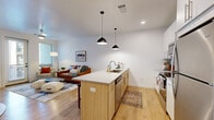 This single-floor residence in West Jordan, Utah, showcases a clean, contemporary design with light-toned luxury vinyl plank flooring and crisp white walls throughout. The open-concept layout connects the kitchen and living area seamlessly, creating an inviting, functional space. The kitchen is well-appointed with stainless steel appliances, including a refrigerator, range, and microwave, complemented by white upper cabinetry with a subway tile backsplash. A peninsula island with a quartz countertop provides additional workspace and casual seating for multiple stools, with two matte black pendant lights overhead adding a modern accent. The living area benefits from natural light through windows and a glass door that opens to a private exterior space. The bedroom offers a calm, understated retreat with windows providing natural light and an adjacent walk-in closet with wire shelving for ample storage. A dedicated workspace area accommodates a desk setup within the bedroom. The bathroom features a wide white vanity with quartz countertop and light wood flooring that carries the cohesive aesthetic throughout. Recessed lighting and a smart lock entry door round out the practical, modern details of this well-designed residence.