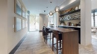 The SkyHouse Nashville leasing office presents a thoughtfully designed, single-floor space that balances professionalism with an inviting atmosphere. Entry is made through glass-paneled doors etched with the SkyHouse Nashville branding, immediately establishing a polished first impression.

The lobby area features soaring ceilings and expansive floor-to-ceiling windows that flood the space with natural light while offering open views of the surrounding streetscape. Neutral-toned, large-format tile flooring runs throughout, lending a cohesive and upscale feel. The seating area is furnished with tufted and leather accent chairs arranged around a contemporary coffee table atop a layered area rug, creating a comfortable yet refined waiting environment. Floor-length textured drapery adds warmth and visual depth.

The reception area is anchored by a striking curved black desk set against a backdrop of curated framed artwork, including an eye-catching abstract piece in teal and gold tones. Recessed lighting highlights the artwork and maintains a well-lit, welcoming corridor leading further into the office.

A bar-height communal table with dark wood bar stools sits beneath a cluster of globe pendant lights, with a branded display shelf behind it. The space flows naturally between zones, creating an efficient and aesthetically cohesive leasing environment throughout.