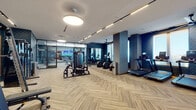 Located at 1301 Spring St NW in Atlanta's Midtown neighborhood, this contemporary fitness center is thoughtfully designed to support a full range of workout disciplines. The space is finished with a striking herringbone-patterned carpet throughout, complemented by deep navy textured accent walls that give the facility a polished, upscale aesthetic.

The main exercise floor is equipped with a comprehensive selection of cardio machines, including treadmills, ellipticals, and stationary bikes, all arranged along natural light-filled windows that offer views of the surrounding cityscape. A dedicated strength training area features a Precor cable machine, adjustable benches, and a full dumbbell rack. Ceiling-mounted track lighting and large round pendant fixtures ensure the space is well-illuminated at all times.

A separate cycling studio houses a row of spinning bikes facing a large wall-mounted television screen, with kettlebells and additional accessories neatly stored along the perimeter. This room features a tray ceiling with circular pendant lighting and a wall of mirrors, enhancing its studio-like atmosphere.

A functional training zone rounds out the facility, offering TRX suspension straps, resistance bands, medicine balls, and a multi-tier storage rack for free weights and accessories. Yoga mats are mounted on a wall rack near the entry door, and full-length framed mirrors throughout aid in form and technique. The overall design balances form and function seamlessly.