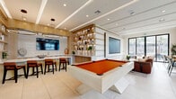 Located at 101 Chamomile Glen in Escondido, CA, this thoughtfully designed single-floor amenity space offers a sophisticated blend of entertainment, relaxation, and social function. The interior is anchored by a striking contemporary pool table with a bold orange felt surface and a sculptural white base, positioned beneath a coffered ceiling with recessed lighting that sets a warm, inviting ambiance. A large projection screen and additional wall-mounted display provide ample entertainment options for gatherings. A built-in shelving unit with integrated backlighting serves as an elegant room divider, displaying curated décor and greenery while delineating the lounge and bar areas. The wet bar features white subway tile backsplash, a sleek countertop, and bar seating with rust-toned leather chairs beneath a statement circular pendant light. The lounge area is generously appointed with plush seating, including a curved cognac sofa accented with vibrant throw pillows, oriented toward expansive floor-to-ceiling windows that flood the space with natural light and offer open views of the surrounding streetscape. A dedicated workspace with counter-height seating lines the window wall, ideal for remote work or casual study. Large-format tile flooring and warm wood accents throughout lend the space a cohesive, modern aesthetic.