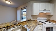 This single-floor residence offers a cohesive and functional layout with warm wood-look flooring flowing consistently throughout. The kitchen is a standout feature, equipped with white cabinetry, granite-style countertops, a stainless steel double sink, a white electric range, and an under-cabinet range hood. A large sliding window above the sink allows natural light to filter in while providing views of the surrounding outdoor greenery. The open layout of the kitchen connects seamlessly to the rest of the living spaces, making the area feel inviting and practical. The living room is generously proportioned, featuring dual windows dressed with vertical blinds that bring in abundant natural light. A ceiling fan with a built-in light fixture adds both comfort and functionality. The bedrooms are well-sized, each outfitted with the same rich wood-look flooring, neutral walls, and vertical blind-covered windows. One bedroom includes an open closet with built-in shelving, providing ample storage. A hallway connects the rooms with ease. The bathroom is cleanly appointed with a full tub-shower combination, a white vanity with granite-style countertops, a large mirror, and wood-look flooring that ties the space together. Textured ceilings are present throughout, adding a classic residential touch to this well-maintained and move-in-ready home.