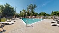 Located in Pikesville, MD, this outdoor pool facility is a well-appointed recreational amenity set within a multi-building residential community. The centerpiece is a generously sized rectangular swimming pool with clear, blue-tinted water, lane markings, and stainless steel entry railings for safe and easy access. The surrounding concrete deck provides ample space for lounging, lined with multiple reclining chaise lounge chairs and patio umbrellas positioned throughout for shade and comfort. A dedicated lifeguard chair is stationed poolside, underscoring the facility's commitment to safety. Bordering the pool on one side is a classic single-story brick pool house featuring a dark shingled roof, decorative trim detailing, and multiple access doors — likely housing restrooms and storage. The entire pool area is enclosed by a combination of wooden privacy fencing and iron security fencing, creating a secure and private atmosphere. Mature trees surrounding the perimeter provide natural shade and a lush green backdrop under open blue skies. The adjacent multi-story brick residential buildings with balconies are visible nearby, connecting the pool area to the broader community. The grounds also feature a well-maintained lawn area, completing this inviting outdoor retreat.