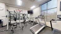 Located at 293 Polifly Rd in Hackensack, NJ, this fitness center offers a thoughtfully organized, single-floor exercise space designed to accommodate a wide range of workout routines. The facility features a clean, modern aesthetic with white walls, a drop-panel ceiling with recessed fluorescent lighting, and durable rubber flooring throughout.

The space is divided into distinct functional zones. The cardio area is outfitted with treadmills, stair climbers, elliptical machines, and stationary bikes, complemented by wall-mounted flat-screen TVs and large windows that allow natural light to filter in. The strength training section includes a comprehensive selection of cable and resistance machines, adjustable benches, and a dedicated free weights area with a full rack of color-coded dumbbells, all set against large wall mirrors that enhance the sense of openness.

Additional zones accommodate functional and specialty training, featuring a heavy punching bag, stability balls, a BOSU ball, and multi-use cable stations. Motivational wall graphics — including the phrase "What the Mind Believes, the Body Achieves" — add an energizing atmosphere to the space. Structural columns are integrated throughout, and glass-panel interior doors provide visibility across the floor. The overall ambiance is bright, clean, and purpose-built for an effective fitness experience.