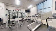 Located at 293 Polifly Rd in Hackensack, NJ, this fitness center offers a thoughtfully organized, single-floor exercise space designed to accommodate a wide range of workout routines. The facility features a clean, modern aesthetic with white walls, a drop-panel ceiling with recessed fluorescent lighting, and durable rubber flooring throughout.

The space is divided into distinct functional zones. The cardio area is outfitted with treadmills, stair climbers, elliptical machines, and stationary bikes, complemented by wall-mounted flat-screen TVs and large windows that allow natural light to filter in. The strength training section includes a comprehensive selection of cable and resistance machines, adjustable benches, and a dedicated free weights area with a full rack of color-coded dumbbells, all set against large wall mirrors that enhance the sense of openness.

Additional zones accommodate functional and specialty training, featuring a heavy punching bag, stability balls, a BOSU ball, and multi-use cable stations. Motivational wall graphics — including the phrase "What the Mind Believes, the Body Achieves" — add an energizing atmosphere to the space. Structural columns are integrated throughout, and glass-panel interior doors provide visibility across the floor. The overall ambiance is bright, clean, and purpose-built for an effective fitness experience.