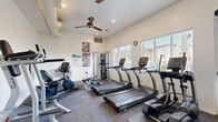 This single-level fitness center at Arbor Park in Vallejo, CA (94590) offers a comprehensive range of exercise equipment within a bright, efficiently organized space. The room features white walls and carpeted flooring, contributing to a clean and functional atmosphere. Natural light filters in through multiple windows fitted with white blinds, providing views of the surrounding community.

The cardio section is anchored by a row of TRUE-brand treadmills and includes elliptical trainers and a stationary cycle, catering to a variety of aerobic workouts. For strength training, the facility houses a Hoist multi-station cable machine, a weight bench, and a dumbbell rack. A large wall mirror enhances the sense of space and allows users to monitor their form during exercise.

Ceiling fans maintain air circulation throughout the room, complementing the HVAC system visible through wall vents. A wall-mounted television provides entertainment during workouts. Posted fitness center rules and exercise reference charts are displayed on the walls. A water fountain is conveniently accessible within the space. Recessed lighting supplements the natural light, ensuring the room remains well-lit throughout the day.