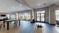 Located at 550 Pitt Greene Connector in Greenville, NC, this single-floor amenity space offers a well-appointed, modern environment designed for both relaxation and productivity. The layout flows seamlessly between several distinct zones, each thoughtfully designed with contemporary finishes throughout.

The main entry welcomes visitors with large floor-to-ceiling windows and glass-paneled doors that flood the space with natural light and offer views of the surrounding exterior. Light wood-look flooring and neutral gray tones establish a clean, cohesive aesthetic throughout.

A spacious social lounge anchors the space, featuring plush white sofas, patterned accent chairs, a pool table, a shuffleboard table, and a distinctive herringbone-tiled accent wall with a mounted flat-screen television. An elegant chandelier adds a refined touch overhead.

Adjacent seating areas include a sunlit nook with deep purple upholstered chairs arranged around a floor-to-ceiling window wall, creating an inviting spot with outdoor courtyard views. A communal high-top table with industrial metal stools offers flexible gathering space near the entrance.

A co-working and business area provides a functional workspace, while a fitness center—accessible through sliding barn doors—is visible from the main corridor. Vending machines and a central service counter round out the practical amenities, making this a comprehensive, community-focused facility.