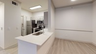 This single-floor unit offers a thoughtfully designed layout that balances functionality and comfort. The kitchen serves as a central highlight, featuring a suite of stainless steel appliances including a refrigerator, electric range, over-the-range microwave, and dishwasher. Gray shaker-style cabinetry pairs with crisp white quartz countertops and a subway tile backsplash, while a peninsula with an integrated sink opens the space to the adjoining living area. Light wood-look flooring runs throughout the kitchen and main living areas, lending a clean, contemporary feel. The bedroom is finished with soft, neutral carpet underfoot and features crown molding, natural light from horizontal blinds-covered windows, and a ceiling fan with integrated lighting for year-round comfort. The full bathroom is outfitted with a tub-shower combination surrounded by white and neutral-toned tile, brushed nickel fixtures, and light wood-look flooring. A connecting hallway with additional cabinetry and a secondary toilet area provides added convenience. Throughout the unit, a neutral color palette of soft whites and warm beiges creates a bright, cohesive ambiance that is move-in ready and easy to personalize.