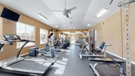 This fitness center offers a spacious, single-room layout designed to support a comprehensive workout experience. The room features vaulted ceilings that lend an open, airy feel, complemented by two ceiling fans that promote air circulation throughout the space. Warm, golden-toned walls are paired with a durable dark rubber floor, creating a practical yet inviting athletic environment.

Natural light streams in generously through multiple windows fitted with blinds, casting bright patterns across the floor and enhancing the overall energy of the room. A glass door at one end further connects the interior to the outdoors, adding to the sense of openness.

The fitness center is well-stocked with a variety of SportsArt-brand cardio equipment, including treadmills, elliptical trainers, and stair climbers, arranged in organized rows along the length of the space. Strength-training equipment, including cable machines and multi-function weight stations, occupies the perimeter. A dedicated free weights area features a dumbbell rack, exercise mats, and a vertical knee raise tower, accompanied by a large wall mirror that enhances spatial depth. Wall-mounted televisions provide entertainment during workouts. The layout ensures ample room to move freely between stations, making this fitness center a functional and comfortable amenity.