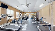 This fitness center offers a spacious, single-room layout designed to support a comprehensive workout experience. The room features vaulted ceilings that lend an open, airy feel, complemented by two ceiling fans that promote air circulation throughout the space. Warm, golden-toned walls are paired with a durable dark rubber floor, creating a practical yet inviting athletic environment.

Natural light streams in generously through multiple windows fitted with blinds, casting bright patterns across the floor and enhancing the overall energy of the room. A glass door at one end further connects the interior to the outdoors, adding to the sense of openness.

The fitness center is well-stocked with a variety of SportsArt-brand cardio equipment, including treadmills, elliptical trainers, and stair climbers, arranged in organized rows along the length of the space. Strength-training equipment, including cable machines and multi-function weight stations, occupies the perimeter. A dedicated free weights area features a dumbbell rack, exercise mats, and a vertical knee raise tower, accompanied by a large wall mirror that enhances spatial depth. Wall-mounted televisions provide entertainment during workouts. The layout ensures ample room to move freely between stations, making this fitness center a functional and comfortable amenity.