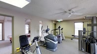 This single-floor fitness space offers a practical and well-equipped environment for cardiovascular exercise. The room features a collection of cardio machines, including elliptical trainers, a treadmill, a stationary bike, and a weight bench, providing a variety of workout options. Natural light filters in through multiple windows, creating a bright and inviting atmosphere, while a ceiling fan overhead ensures adequate air circulation during workouts. The textured ceiling adds a classic touch to the space. A skylight further enhances the room's brightness. Through the interior windows, an adjacent room is visible, appearing to contain a billiards table, suggesting this fitness area is part of a larger recreational facility. The neutral-toned carpet and light-colored walls contribute to a clean, functional aesthetic. An emergency exit sign is visible near a doorway, indicating the space meets safety standards. Overall, this fitness room balances practicality and comfort, making it a convenient and accessible space for regular exercise.