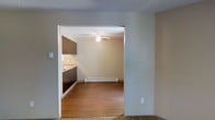 This single-floor space offers a straightforward and functional layout designed for comfortable everyday living. The interior is characterized by warm hardwood flooring that flows through the main living and kitchen areas, creating a cohesive and inviting atmosphere. A wide open archway connects the kitchen to the living area, promoting an open-concept feel that enhances the sense of space. The kitchen is fitted with dark-toned cabinetry providing ample storage, a light-colored countertop, and a sink positioned along the wall — offering a practical and efficient workspace. The living area is equipped with a ceiling fan, which aids in air circulation and adds a functional overhead feature. A baseboard heater runs along the far wall, ensuring consistent warmth throughout the space. The ceilings feature a textured finish, adding subtle visual depth to the rooms. The neutral wall tones throughout create a clean, versatile backdrop that suits a variety of interior styles. Overall, this space combines practical amenities with a simple, well-connected layout that makes efficient use of its single-floor footprint.