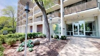 This multi-story residential building presents a welcoming and well-appointed exterior that reflects thoughtful design and upkeep. The building's facade features a classic mid-century architectural style, characterized by clean horizontal lines, textured brick accents, and rows of private balconies that extend across multiple floors — offering residents outdoor space with open views.

The main entrance is accessible via a broad, paved walkway framed by carefully maintained landscaping. Mature trees provide natural shade and greenery, complementing the neatly trimmed shrubs and ornamental plantings that line the grounds. Natural boulders are incorporated into the landscaping, adding an organic, grounded aesthetic to the exterior environment.

Sleek, modern lampposts are positioned along the pathway, ensuring the entrance remains well-lit and inviting at all hours. The entrance itself features floor-to-ceiling glass doors and expansive glazing, creating a bright and transparent threshold that connects the outdoor landscaped setting to the interior lobby. The glass-fronted entry allows natural light to flow freely into the building's common areas.

Overall, the exterior conveys a sense of established, comfortable residential living, where architectural structure and natural elements work in harmony to create an attractive and functional entryway.
