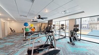 Located at 101 Viva Bene Circle in Saint Peters, Missouri, this fitness and wellness center offers a thoughtfully designed, single-floor layout encompassing a generously sized exercise room, a multipurpose living/activity room, and a dedicated bedroom or rest space. The exercise room is the centerpiece of the facility, outfitted with a comprehensive array of cardio equipment including treadmills, ellipticals, stair climbers, and Concept2 rowing machines. A full selection of free weights, dumbbells, kettlebells, and stability balls in vibrant colors rounds out the strength training offerings. The bold "Purposefully Active" wall mural sets an motivating tone throughout the space. Distinctive teal and gray chevron-patterned carpet adds a modern, energetic aesthetic underfoot, while multiple ceiling fans ensure comfort during workouts. Large floor-to-ceiling windows flood the cardio area with natural light and provide views of the surrounding outdoor areas. Adjacent to the exercise room is a bright, open studio space featuring smooth hardwood-style flooring, a full wall of mirrors ideal for group classes or individual training, and vibrant artwork that adds personality. A small kitchenette and seating area provide functional support spaces. The overall ambiance is clean, modern, and purpose-driven.