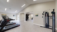 This single-floor fitness facility offers a thoughtfully designed workout environment with a clean, modern aesthetic. The main exercise area is defined by soaring vaulted ceilings and sleek linear LED lighting, creating a bright and energizing atmosphere. Light-toned luxury vinyl plank flooring runs throughout, complementing the crisp white walls and white baseboards for a polished, professional look.

The space is divided into distinct functional zones. The primary cardio area houses multiple treadmills and an elliptical trainer, positioned alongside windows that allow natural light to flow in. A secondary zone, partially separated by a structural column and open pass-through, features a Peloton-style spin bike, a weight bench, a dumbbell rack, and a flat-screen TV mounted on a striking shiplap accent wall — ideal for guided training sessions.

An additional strength training area includes a multi-functional cable machine, providing a comprehensive resistance workout option. Coastal-themed artwork is displayed on the walls throughout, adding a calming, motivational ambiance to the space. A door connects the fitness area to an adjacent interior space, further enhancing the facility's accessibility and flow. Overall, this fitness space balances functionality with inviting design, making it well-suited for a full range of workout routines.
