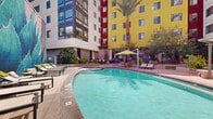 Located in the heart of downtown Tucson at 350 E Congress Street, this outdoor courtyard amenity level is a dynamic, well-appointed space designed for both relaxation and socializing. The centerpiece is a generously sized resort-style swimming pool accompanied by a spa, surrounded by lounge chairs and shade umbrellas, creating an inviting retreat under the Arizona sun. Tall palm trees and mature shade trees are integrated throughout, offering natural relief from the heat while adding a lush, tropical ambiance. The courtyard is framed by the bold, multi-story building façade featuring striking yellow, red, and white exterior panels that lend the space a vibrant, contemporary urban character. Raised concrete planters filled with native desert vegetation — including agave and other drought-tolerant plants — define pathways and seating areas across the space. A dedicated green lawn area provides an open, flexible gathering zone, complemented by concrete benches for casual seating. A tiled, built-in bench nook offers a more intimate outdoor retreat. The outdoor kitchen area is a standout feature, equipped with stainless steel grills, a granite-topped bar counter, and bar-height seating — ideal for outdoor dining and entertaining. A vibrant mural depicting a desert agave plant adds an artistic focal point to the pool area, reinforcing the local Sonoran Desert identity of this thoughtfully designed urban outdoor space.