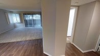 This single-story space presents a practical and straightforward layout, with design elements that emphasize functionality. The main living area is defined by a clear transition between rich, dark hardwood flooring near the entry and plush carpeting that extends through the primary living space. This combination provides both visual contrast and practical comfort underfoot.

The living area is well-lit by natural light streaming through a large sliding glass door fitted with vertical blinds, which allows for adjustable light control and provides direct outdoor access. A side window further supplements natural lighting, contributing to a bright and open atmosphere. The neutral gray wall tones throughout create a clean, understated backdrop that suits a variety of interior styles.

A textured popcorn ceiling spans the space, consistent with classic residential construction. Structural columns partially divide areas of the interior, offering a subtle sense of separation between spaces without fully enclosing them. A hallway visible from the main area leads to additional rooms, with the hardwood flooring continuing into this transitional corridor. White baseboards provide a crisp finish along the walls throughout the space.