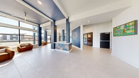 This contemporary lobby and common area presents a striking first impression through its thoughtful architectural design and cohesive color palette. Bold navy-blue columns and ceiling accents contrast sharply against crisp white walls and light-toned large-format tile flooring, creating a visually dynamic yet polished environment. The space benefits from an abundance of natural light, courtesy of expansive floor-to-ceiling windows and black-framed glass walls that wrap around multiple corners, offering panoramic outdoor views and inviting sunlight deep into the interior.

A central reception desk serves as a focal point, clad in a vibrant, abstract multicolored panel that doubles as a work of art. A decorative globe-inspired wall sculpture mounted on a deep navy accent wall reinforces the space's modern aesthetic. The open layout accommodates distinct functional zones, including comfortable lounge seating areas with rounded cognac-toned chairs positioned near the windowed corners, creating intimate gathering spots bathed in natural light.

A dedicated event room, accessible through black-framed glass double doors, extends the usability of the space. Throughout, recessed lighting and statement pendant fixtures complement the natural illumination. Artwork and potted greenery add warmth and personality, balancing the clean, contemporary lines of the architecture. The overall ambiance is bright, welcoming, and professionally designed.