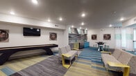 This single-floor community lounge is a well-appointed social space designed for gathering, recreation, and relaxation. The interior is unified by a striking patchwork carpet in bold shades of teal, yellow, and gray, which adds energy and visual interest throughout the open layout.

The game and entertainment zone anchors one end of the space, featuring a full-length shuffleboard table, a wall-mounted flat-screen television, and decorative wall art with a retro-music theme. Comfortable patterned seating with modern yellow accent side tables is arranged to encourage casual socializing in this area.

Adjacent to the lounge is a fully equipped kitchenette, distinguished by its colorful cabinetry in teal, yellow, and gray tones, complemented by a textured stone mosaic backsplash and open display shelving. Stainless steel appliances add a contemporary touch, while a round high-top table with teal bar stools provides a casual dining or gathering spot.

A secondary seating area with additional wall-mounted televisions and a media console offers a quieter retreat within the same space. Restroom facilities are conveniently located nearby, including an ADA-accessible option. Recessed lighting throughout ensures a bright, welcoming ambiance, and large windows allow natural light to filter in, enhancing the overall open and inviting character of the lounge.