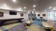 This single-floor community lounge is a well-appointed social space designed for gathering, recreation, and relaxation. The interior is unified by a striking patchwork carpet in bold shades of teal, yellow, and gray, which adds energy and visual interest throughout the open layout.

The game and entertainment zone anchors one end of the space, featuring a full-length shuffleboard table, a wall-mounted flat-screen television, and decorative wall art with a retro-music theme. Comfortable patterned seating with modern yellow accent side tables is arranged to encourage casual socializing in this area.

Adjacent to the lounge is a fully equipped kitchenette, distinguished by its colorful cabinetry in teal, yellow, and gray tones, complemented by a textured stone mosaic backsplash and open display shelving. Stainless steel appliances add a contemporary touch, while a round high-top table with teal bar stools provides a casual dining or gathering spot.

A secondary seating area with additional wall-mounted televisions and a media console offers a quieter retreat within the same space. Restroom facilities are conveniently located nearby, including an ADA-accessible option. Recessed lighting throughout ensures a bright, welcoming ambiance, and large windows allow natural light to filter in, enhancing the overall open and inviting character of the lounge.