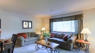 This single-floor space centers around a welcoming living room designed with comfort and functionality in mind. The room features neutral-toned walls that complement the warm, beige carpeting underfoot, creating a cohesive and inviting atmosphere throughout. Two generously sized sofas — one a classic tan fabric style and the other a larger grey sectional — are arranged to encourage conversation, each accented with decorative throw pillows in deep red and muted tones. A large window dressed with vertical blinds and a dark valance allows abundant natural light to fill the room, enhancing its open and airy feel. A wrought-iron and wood coffee table serves as a stylish focal point at the center of the seating arrangement, complemented by a matching side table with a traditional table lamp that adds warmth to the space. A framed mirror on the wall adds depth and reflects light effectively. The textured ceiling and clean wall finishes contribute to the room's polished yet relaxed character, making this a well-balanced living space suitable for both everyday use and casual entertaining.