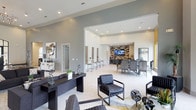 This sophisticated common area is designed to impress, combining modern aesthetics with a welcoming, functional layout across a single floor. The grand entrance lobby sets the tone immediately, boasting soaring ceilings, large arched windows that flood the space with natural light, and views of a pool and lush greenery beyond. Elegant globe chandeliers and recessed lighting complement the crisp white walls and large-format tile flooring throughout.

The main lounge area features multiple seating arrangements with sleek black leather chairs, dark wood accent tables, and curated wall art, creating an inviting yet refined atmosphere. A central reception desk anchors the space, surrounded by open sightlines that connect seamlessly to the adjacent dining and entertainment zones.

The dining area is furnished with contemporary high-back chairs around a long table, positioned near a striking feature wall clad in a bold mosaic tile backsplash. A flat-screen television is mounted above dark cabinetry that houses dual wine coolers and ample counter space, ideal for gatherings and social events.

A casual café-style nook with bistro-height tables and patterned upholstered stools adds a relaxed, social dimension to the space. The kitchenette is finished with quartz countertops, dark cabinetry, a stainless steel sink, and a window overlooking the outdoor surroundings, providing both practicality and charm.