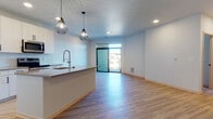 Located at 6414 Bridge Rd in Monona, WI, this single-floor unit presents a thoughtfully designed open-concept layout with a clean, contemporary aesthetic. Light-toned hardwood-style flooring flows consistently throughout the space, complementing the bright white walls and recessed lighting that keep the interior feeling fresh and well-illuminated.

The kitchen and living area share an open, connected layout. The kitchen is outfitted with white cabinetry, stainless steel appliances including a refrigerator, range, and microwave, and a tile backsplash. A peninsula with a quartz-style countertop provides additional workspace and casual seating. Two geometric pendant lights hang above, adding a modern design accent. A floor-to-ceiling sliding glass door at the far end of the living area fills the space with natural light and provides direct access to an outdoor balcony.

The bedroom features a built-in closet with sliding light wood doors and wire shelving for organized storage. The bathroom is finished with a wide vanity featuring textured cabinetry, black hardware, a large mirror, and a tub-shower combination with open shelving alongside it. A small interior window connects the kitchen to an adjacent hallway, adding an architectural detail that enhances the sense of openness throughout the unit.