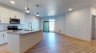 Located at 6414 Bridge Rd in Monona, WI, this single-floor unit presents a thoughtfully designed open-concept layout with a clean, contemporary aesthetic. Light-toned hardwood-style flooring flows consistently throughout the space, complementing the bright white walls and recessed lighting that keep the interior feeling fresh and well-illuminated.

The kitchen and living area share an open, connected layout. The kitchen is outfitted with white cabinetry, stainless steel appliances including a refrigerator, range, and microwave, and a tile backsplash. A peninsula with a quartz-style countertop provides additional workspace and casual seating. Two geometric pendant lights hang above, adding a modern design accent. A floor-to-ceiling sliding glass door at the far end of the living area fills the space with natural light and provides direct access to an outdoor balcony.

The bedroom features a built-in closet with sliding light wood doors and wire shelving for organized storage. The bathroom is finished with a wide vanity featuring textured cabinetry, black hardware, a large mirror, and a tub-shower combination with open shelving alongside it. A small interior window connects the kitchen to an adjacent hallway, adding an architectural detail that enhances the sense of openness throughout the unit.
