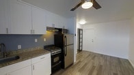 This single-floor unit offers a thoughtfully designed layout with a clean, modern aesthetic. The kitchen is a functional highlight, featuring white shaker-style cabinetry that extends to the ceiling for ample storage, complemented by granite countertops with a subtle speckled finish. Stainless steel appliances include a full-size refrigerator, an electric range with oven, and a microwave, all positioned efficiently along one wall. An under-cabinet range hood adds both practicality and a streamlined look. The soft blue-gray wall color provides a calming backdrop that contrasts crisply with the white cabinetry. Wood-look plank flooring runs continuously through the kitchen and into the adjoining areas, adding warmth and visual cohesion throughout the space. A ceiling fan with an integrated light fixture provides both air circulation and ambient lighting. The entryway is visible from the kitchen, featuring a solid front door and a sleek sliding frosted-glass interior door, adding a modern design element. The open sightlines between the kitchen and entry create an airy, connected feel within this efficiently designed single-level unit.