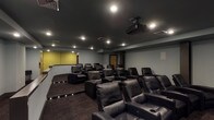 The Phase II Theater is a purpose-built, single-floor screening room designed to deliver an immersive cinematic experience. The main auditorium is defined by its dark, dramatic aesthetic, with dark-painted ceilings and walls that minimize light reflection and enhance viewing conditions. Seating is arranged in a tiered configuration, with rows of oversized black leather reclining chairs equipped with built-in cup holders, providing both comfort and clear sightlines to the screen from every position. Step lighting along the tiered risers ensures safe navigation in low-light conditions.

A ceiling-mounted projector is centrally positioned to deliver a clear, high-quality image onto the large, black-bordered projection screen mounted on the front wall. Multiple in-wall speakers are strategically placed throughout the room to support a surround sound audio setup. Recessed ceiling lighting provides ambient illumination when the theater is not in use.

Adjacent to the main screening room is a rear lobby or transition area, featuring gray walls, dark baseboards, dark carpet tile flooring, and a granite-topped cabinetry unit, offering a functional space for gathering before or after screenings.