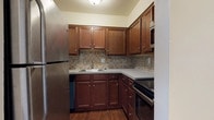 This single-floor residence offers a practical and comfortable layout designed for everyday living. The kitchen is a standout feature, showcasing rich, warm-toned cabinetry with brushed nickel hardware, grey countertops, and a classic tile backsplash in a neutral mosaic pattern. Stainless steel appliances — including a refrigerator, range, built-in microwave, and dishwasher — complete the space, which is finished with durable hardwood-style flooring.

The open living area is generously proportioned, featuring the same hardwood-style flooring, clean white walls, and vertical blinds that allow natural light to filter in through a sliding door. A closet and wall-mounted thermostat add to the functionality of this main living space.

The bedroom is a calm, carpeted retreat with a wide window that brings in natural light. It connects directly to a utility closet housing a stacked washer and dryer, as well as a dedicated bathroom. The bathroom is clean and bright, featuring a full tub and shower combo with built-in shelving, white surround walls, and hardwood-style flooring that ties the space together cohesively. The overall ambiance is neutral, tidy, and move-in ready.