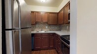 This single-floor residence offers a practical and comfortable layout designed for everyday living. The kitchen is a standout feature, showcasing rich, warm-toned cabinetry with brushed nickel hardware, grey countertops, and a classic tile backsplash in a neutral mosaic pattern. Stainless steel appliances — including a refrigerator, range, built-in microwave, and dishwasher — complete the space, which is finished with durable hardwood-style flooring.

The open living area is generously proportioned, featuring the same hardwood-style flooring, clean white walls, and vertical blinds that allow natural light to filter in through a sliding door. A closet and wall-mounted thermostat add to the functionality of this main living space.

The bedroom is a calm, carpeted retreat with a wide window that brings in natural light. It connects directly to a utility closet housing a stacked washer and dryer, as well as a dedicated bathroom. The bathroom is clean and bright, featuring a full tub and shower combo with built-in shelving, white surround walls, and hardwood-style flooring that ties the space together cohesively. The overall ambiance is neutral, tidy, and move-in ready.