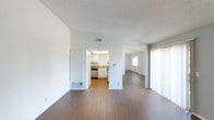 This single-floor residence offers a clean, open layout designed for functional living. The main living area is spacious and bright, featuring warm hardwood-style flooring that extends seamlessly throughout the space, creating a cohesive and inviting atmosphere. Crisp white walls and a textured ceiling complement the light-toned interior, while a large window dressed with sheer white curtains allows natural light to filter in softly.

The kitchen is situated just off the main living area and is visible through an open passageway, offering white cabinetry, a stainless steel dishwasher, and a sink beneath overhead fluorescent lighting. The layout allows for an easy flow between the kitchen and living spaces, making the area practical for everyday use.

A hallway connects to an additional room, which also benefits from natural light through a window and mirrors that enhance the sense of openness. Built-in storage or closet space is incorporated into the living area, adding to the unit's practical design. Wall-mounted HVAC vents provide climate control throughout the space. Overall, the residence presents a straightforward, well-maintained interior with a neutral palette that offers a versatile and comfortable living environment.