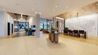 This expansive amenity floor offers a comprehensive and thoughtfully designed collection of spaces unified by a modern aesthetic that blends warm wood accents, large-format tile flooring, and sophisticated finishes throughout. The grand lobby serves as the central hub, featuring striking cylindrical concrete columns, recessed lighting, an artistic sculptural console table, and curated wall art — creating a welcoming and polished first impression. Floor-to-ceiling glass walls and windows invite abundant natural light and provide seamless views to an outdoor courtyard, where a wood-pergola terrace and lush greenery extend the living environment beyond the interior. A dedicated fitness center is well-equipped with cardio machines, free weights, a functional training rig with TRX suspension systems, and full-length mirrors, all set against large windows with outdoor views. The co-working and meeting areas feature elegant steel-framed glass partitions enclosing private conference rooms with designer pendant lighting, ideal for professional use. A stylishly appointed kitchenette offers a dark marble countertop, metallic tile backsplash, and open shelving. The resident lounge area showcases wide-plank hardwood flooring, a sculptural chandelier, plush seating, and an adjacent entertainment room, making the entire floor a complete, resort-caliber amenity experience.