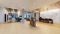This expansive amenity floor offers a comprehensive and thoughtfully designed collection of spaces unified by a modern aesthetic that blends warm wood accents, large-format tile flooring, and sophisticated finishes throughout. The grand lobby serves as the central hub, featuring striking cylindrical concrete columns, recessed lighting, an artistic sculptural console table, and curated wall art — creating a welcoming and polished first impression. Floor-to-ceiling glass walls and windows invite abundant natural light and provide seamless views to an outdoor courtyard, where a wood-pergola terrace and lush greenery extend the living environment beyond the interior. A dedicated fitness center is well-equipped with cardio machines, free weights, a functional training rig with TRX suspension systems, and full-length mirrors, all set against large windows with outdoor views. The co-working and meeting areas feature elegant steel-framed glass partitions enclosing private conference rooms with designer pendant lighting, ideal for professional use. A stylishly appointed kitchenette offers a dark marble countertop, metallic tile backsplash, and open shelving. The resident lounge area showcases wide-plank hardwood flooring, a sculptural chandelier, plush seating, and an adjacent entertainment room, making the entire floor a complete, resort-caliber amenity experience.
