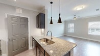 This two-story residence offers a smart and functional layout spread across two levels, combining modern finishes with practical living spaces. The main living level features an open-concept kitchen and living room, where a granite-topped kitchen island with an undermount sink takes center stage, illuminated by a pair of sleek pendant lights. Dark espresso cabinetry provides rich contrast against light-toned walls, while stainless steel appliances — including a refrigerator, range, and built-in microwave — complete the kitchen. A tile backsplash and track lighting add refined detail. The living area flows naturally from the kitchen, bathed in natural light from a window with blinds and finished with wood-look flooring and crown molding throughout. A dedicated laundry room houses a full-size washer and dryer, adding everyday convenience. The upper level hosts the spacious primary bedroom, featuring plush carpeting, crown molding, a ceiling fan, and a large window that welcomes natural light. An adjacent closet provides ample storage. A full bathroom and additional closet space round out the upper floor. The ground level includes a private, attached garage offering secure parking and extra storage. Neutral tones, crown molding, and consistent finishes throughout create a cohesive and polished ambiance.