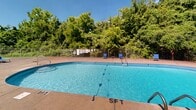 Located at 415 W Northfield Blvd in Murfreesboro, Tennessee, this outdoor poolside amenity area offers a well-appointed communal retreat designed for relaxation and recreation. The centerpiece of the space is a generously sized freeform swimming pool with clearly marked depth zones, including a shallow end and a five-foot section, separated by a floating lane divider. The pool's vivid turquoise water is complemented by a wide, open concrete sundeck that provides ample room for lounging. Multiple blue lounge chairs are arranged throughout the deck, offering plenty of seating options for residents to enjoy the outdoors. The area is enclosed by a combination of ornamental iron fencing with sturdy brick pillar accents and a chain-link perimeter fence, ensuring both security and an open, airy feel. A compact brick utility or restroom structure is situated near the gated entrance, adding functional convenience to the space. The surrounding landscape features mature, leafy trees that provide natural shade and a lush green backdrop, enhancing the overall atmosphere. A ping pong table near the entrance adds an element of recreational variety. The space benefits from open sky views and abundant natural light, making it an inviting outdoor destination throughout the warmer months.