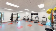 This fitness room at 900 Farmington Ave in West Hartford, Connecticut, offers a comprehensive exercise environment designed to support a wide range of workout routines. The space features a clean, modern aesthetic with crisp white walls, a drop-panel ceiling with recessed fluorescent lighting, and a durable grey floor accented by bold red square tile insets that add a dynamic visual element throughout. Natural light filters in through multiple windows, complementing the bright overhead lighting to create an energizing atmosphere. The room is well-stocked with cardio equipment, including treadmills, elliptical trainers, and a stationary bike, all positioned along the walls to maximize open floor space. A wall-mounted flat-screen television provides entertainment during workouts. Strength training is supported by a cable machine system, a power rack with an adjustable bench, and a full dumbbell rack. Functional training accessories are also well-represented, with a dedicated storage rack holding medicine balls, resistance bands, and stability balls in various sizes. A wall-mounted water fountain provides added convenience. Two interior doors offer access to adjacent areas, making this fitness room a practical and fully integrated wellness facility.
