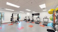 This fitness room at 900 Farmington Ave in West Hartford, Connecticut, offers a comprehensive exercise environment designed to support a wide range of workout routines. The space features a clean, modern aesthetic with crisp white walls, a drop-panel ceiling with recessed fluorescent lighting, and a durable grey floor accented by bold red square tile insets that add a dynamic visual element throughout. Natural light filters in through multiple windows, complementing the bright overhead lighting to create an energizing atmosphere. The room is well-stocked with cardio equipment, including treadmills, elliptical trainers, and a stationary bike, all positioned along the walls to maximize open floor space. A wall-mounted flat-screen television provides entertainment during workouts. Strength training is supported by a cable machine system, a power rack with an adjustable bench, and a full dumbbell rack. Functional training accessories are also well-represented, with a dedicated storage rack holding medicine balls, resistance bands, and stability balls in various sizes. A wall-mounted water fountain provides added convenience. Two interior doors offer access to adjacent areas, making this fitness room a practical and fully integrated wellness facility.