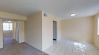 This single-floor space presents a clean, functional layout with a seamless transition between areas. The main living area is finished with light-colored carpet, while an adjacent section features white square tile flooring that reflects natural and artificial light, contributing to a bright, open feel. The walls are painted in a soft, warm neutral tone, creating a calm and understated ambiance throughout.

A partial dividing wall separates the carpeted and tiled sections, adding a subtle sense of spatial definition without fully enclosing the areas. A doorway visible from the main space leads to what appears to be a utility or laundry closet, housing a stacked or side-by-side appliance setup — a practical feature for everyday living.

Ceiling-mounted lighting provides consistent illumination across the space, complemented by HVAC vents integrated into both the ceiling and walls for climate control. White baseboards trim the perimeter of the room, providing a clean contrast against the neutral walls. The textured ceiling adds a modest architectural detail overhead. Overall, the space offers a straightforward, well-maintained interior suited for comfortable day-to-day use.