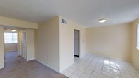 This single-floor space presents a clean, functional layout with a seamless transition between areas. The main living area is finished with light-colored carpet, while an adjacent section features white square tile flooring that reflects natural and artificial light, contributing to a bright, open feel. The walls are painted in a soft, warm neutral tone, creating a calm and understated ambiance throughout.

A partial dividing wall separates the carpeted and tiled sections, adding a subtle sense of spatial definition without fully enclosing the areas. A doorway visible from the main space leads to what appears to be a utility or laundry closet, housing a stacked or side-by-side appliance setup — a practical feature for everyday living.

Ceiling-mounted lighting provides consistent illumination across the space, complemented by HVAC vents integrated into both the ceiling and walls for climate control. White baseboards trim the perimeter of the room, providing a clean contrast against the neutral walls. The textured ceiling adds a modest architectural detail overhead. Overall, the space offers a straightforward, well-maintained interior suited for comfortable day-to-day use.