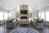 This single-floor activity center is designed to serve as a welcoming communal gathering space, combining functionality with a warm, inviting aesthetic throughout. The centerpiece of the lounge area is a dramatic floor-to-ceiling stacked stone fireplace, flanked by built-in shelving units and topped with a wall-mounted flat-screen television. Soaring vaulted ceilings amplify the sense of openness, while rich hardwood-style flooring runs seamlessly throughout the space. Two plush sofas arranged around a glass coffee table create a comfortable seating area, and large windows invite an abundance of natural light. Adjacent to the lounge, a casual dining area features several high-top granite tables paired with swivel bar stools, making it ideal for informal gatherings. Colorful abstract artwork adorns the walls, and an "Activity Center Rules" display is prominently posted. The fully equipped kitchen boasts crisp white cabinetry, granite countertops, a mosaic tile backsplash, stainless steel appliances including a French door refrigerator and dishwasher, and a central island with additional bar seating. A soft drink station adds a convenient amenity for guests. Recessed lighting throughout ensures a bright, clean atmosphere across every area of this thoughtfully designed community space.