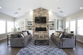 This single-floor activity center is designed to serve as a welcoming communal gathering space, combining functionality with a warm, inviting aesthetic throughout. The centerpiece of the lounge area is a dramatic floor-to-ceiling stacked stone fireplace, flanked by built-in shelving units and topped with a wall-mounted flat-screen television. Soaring vaulted ceilings amplify the sense of openness, while rich hardwood-style flooring runs seamlessly throughout the space. Two plush sofas arranged around a glass coffee table create a comfortable seating area, and large windows invite an abundance of natural light. Adjacent to the lounge, a casual dining area features several high-top granite tables paired with swivel bar stools, making it ideal for informal gatherings. Colorful abstract artwork adorns the walls, and an "Activity Center Rules" display is prominently posted. The fully equipped kitchen boasts crisp white cabinetry, granite countertops, a mosaic tile backsplash, stainless steel appliances including a French door refrigerator and dishwasher, and a central island with additional bar seating. A soft drink station adds a convenient amenity for guests. Recessed lighting throughout ensures a bright, clean atmosphere across every area of this thoughtfully designed community space.