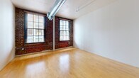 This single-floor loft-style unit blends industrial character with modern finishes to create a functional and visually compelling living space. The main living area is defined by its exposed red brick accent walls, large multi-pane windows that invite abundant natural light, and a prominent exposed spiral HVAC duct running across the ceiling — hallmarks of classic industrial loft design. Light-toned hardwood flooring runs throughout the space, adding warmth and continuity.

The open floor plan flows seamlessly from the main living area into the kitchen and ancillary spaces. The kitchen is well-appointed with a two-tone cabinet scheme featuring rich dark lower cabinetry contrasted against white upper cabinets, complemented by a matching dark refrigerator and dishwasher. A central column anchors the kitchen area, adding architectural interest. Recessed lighting and track lighting fixtures illuminate the kitchen and hallway areas effectively.

A dedicated bathroom features a clean, minimal layout with neutral tones and tile flooring. Additional interior doors along the hallway suggest closet or storage spaces, contributing to the unit's practicality. The industrial-modern aesthetic throughout — from the raw brick to the clean white walls and polished floors — gives this space a distinctive urban character suited for a variety of uses.