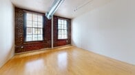 This single-floor loft-style unit blends industrial character with modern finishes to create a functional and visually compelling living space. The main living area is defined by its exposed red brick accent walls, large multi-pane windows that invite abundant natural light, and a prominent exposed spiral HVAC duct running across the ceiling — hallmarks of classic industrial loft design. Light-toned hardwood flooring runs throughout the space, adding warmth and continuity.

The open floor plan flows seamlessly from the main living area into the kitchen and ancillary spaces. The kitchen is well-appointed with a two-tone cabinet scheme featuring rich dark lower cabinetry contrasted against white upper cabinets, complemented by a matching dark refrigerator and dishwasher. A central column anchors the kitchen area, adding architectural interest. Recessed lighting and track lighting fixtures illuminate the kitchen and hallway areas effectively.

A dedicated bathroom features a clean, minimal layout with neutral tones and tile flooring. Additional interior doors along the hallway suggest closet or storage spaces, contributing to the unit's practicality. The industrial-modern aesthetic throughout — from the raw brick to the clean white walls and polished floors — gives this space a distinctive urban character suited for a variety of uses.