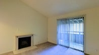 This single-floor space presents a straightforward and functional living area centered around two notable features: a fireplace and a sliding glass door. The fireplace, positioned along one wall, features a tile surround and a simple white mantel, serving as a natural focal point within the room. Its classic design adds a touch of warmth and character to the otherwise neutral interior.

Opposite the fireplace, a full-width sliding glass door dominates the exterior wall, dressed with vertical blinds that allow for adjustable light control. The door provides direct access to an outdoor area and draws in natural light, brightening the space considerably. The interplay between the blinds and incoming light creates a clean, airy atmosphere throughout the room.

The walls are finished in a light, neutral tone, complementing the beige carpeted flooring that extends across the room. The carpet provides a soft underfoot experience and contributes to the overall understated aesthetic. The ceiling features a subtle textured finish and follows a standard height, maintaining a comfortable sense of proportion within the room. Overall, the space offers a clean, move-in-ready layout suited for practical everyday living.