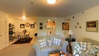 This single-floor residence offers a warm and inviting atmosphere with a thoughtfully designed open-concept layout that connects the main living spaces seamlessly. The living room serves as the heart of the home, featuring plush, comfortable seating in soft sage tones accented with classic plaid throw pillows, lending the space a relaxed, cottage-inspired charm. Ceiling-mounted lighting provides a soft, ambient glow throughout, complementing the neutral wall tones and light carpeting underfoot.

The living area flows naturally into a dedicated dining space, where a traditional wooden dining table and chairs sit atop a rich area rug, creating a defined and intimate setting for meals. Decorative wall art, a classic wall clock, and tasteful accessories throughout add personal character and warmth to the home.

Just off the main living area, the kitchen is easily visible through a central opening, revealing light wood cabinetry, white appliances including a refrigerator and microwave, and a clean, functional layout. The kitchen's efficient design ensures everything is within easy reach.

Overall, this residence exudes a cozy, homey ambiance with its cohesive décor, soft lighting, and well-connected living spaces — ideal for comfortable everyday living.
